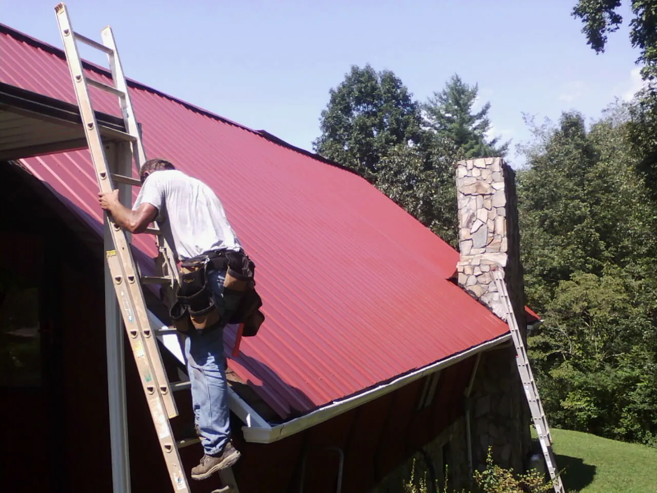 Roofer on a ladder beside a red metal roof and stone chimney — Soffit & Fascia Repair by Roofing Brothers in Nine Mile Falls